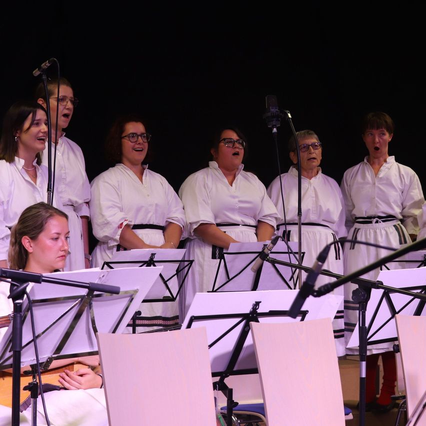 Ein Chor von Frauen in weißen Gewändern singt auf der Bühne mit Mikrofonen und Notenblättern vor ihnen.