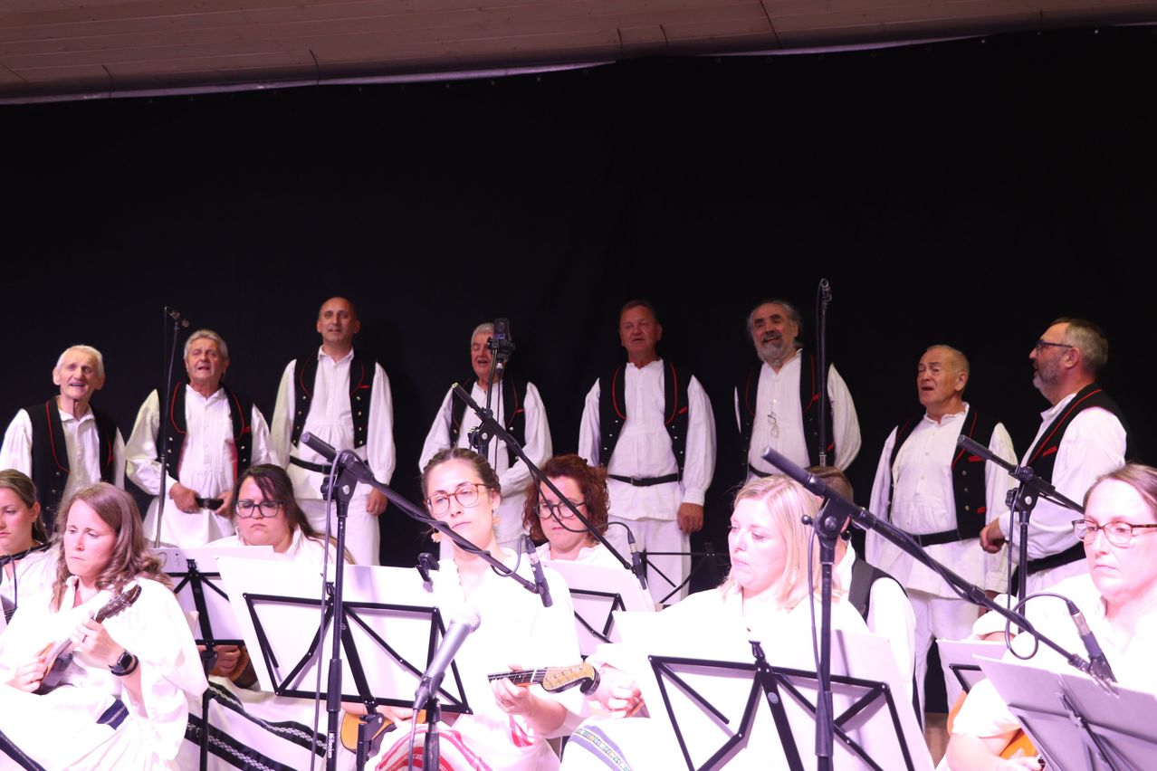 Eine Gruppe von Menschen in weißer traditioneller Kleidung tritt auf der Bühne mit Musikinstrumenten und Mikrofonen vor einem schwarzen Hintergrund auf.