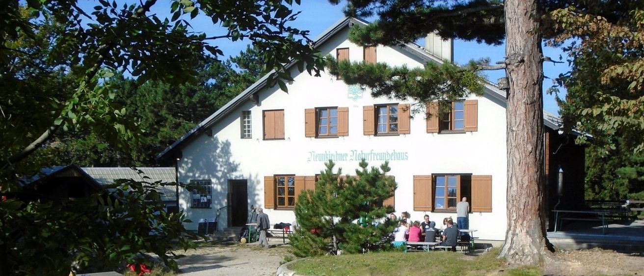 Ein weißes Gebäude mit braunen Fensterläden, möglicherweise ein Gästehaus, hat mehrere Menschen davor versammelt. Sie sitzen auf einer Bank unter einem Baum, andere stehen in der Nähe. Die Szene ist im Freien mit Holzbänken und einer Steinmauer.