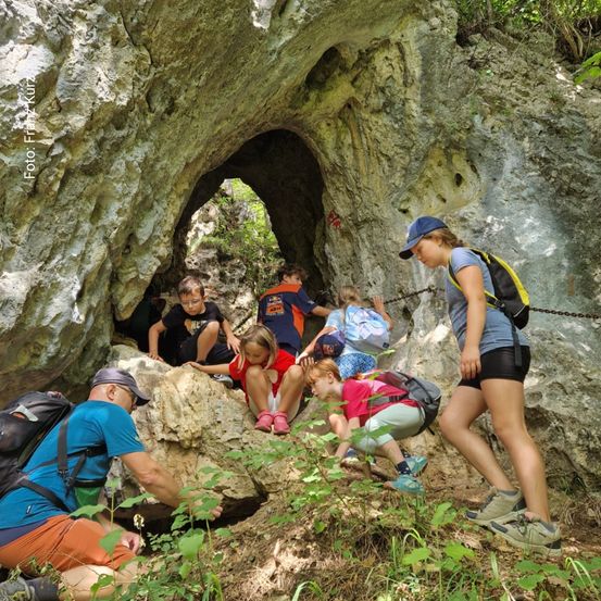 Eine Gruppe von Kindern und Erwachsenen wandert auf einem steilen felsigen Gelände, einige klettern und andere sitzen auf Felsen. Die Umgebung ist draußen mit üppiger Vegetation.