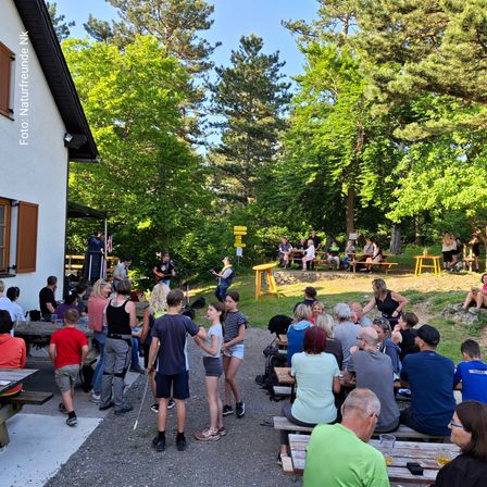 Eine Gruppe von Menschen versammelt sich in einem Park, einige sitzen auf Bänken, andere stehen, mit einem Gebäude und üppiger Grünfläche im Hintergrund. Eine Person spielt Gitarre auf einer Bühne, und andere genießen Getränke an Picknicktischen.