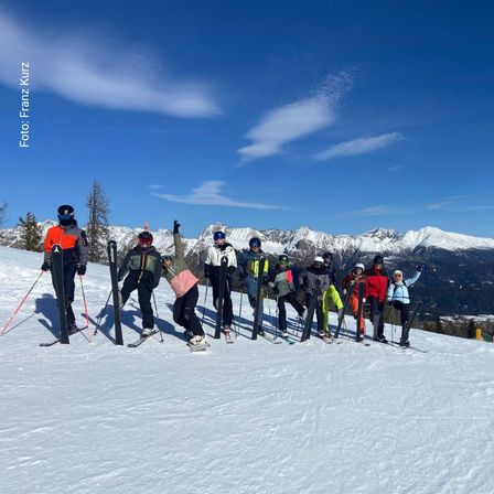 Eine Gruppe von Skifahrern posiert für ein Foto auf einem verschneiten Berg. Sie tragen Winterkleidung und Helme, und der Himmel ist klar mit einigen Wolken. Der Schnee ist weiß und makellos, und die Berge sind schneebedeckt.
