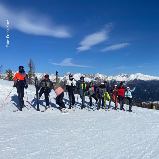Eine Gruppe von Skifahrern posiert für ein Foto auf einem verschneiten Berg. Sie tragen Winterkleidung und Helme, und der Himmel ist klar mit einigen Wolken. Der Schnee ist weiß und makellos, und die Berge sind schneebedeckt.