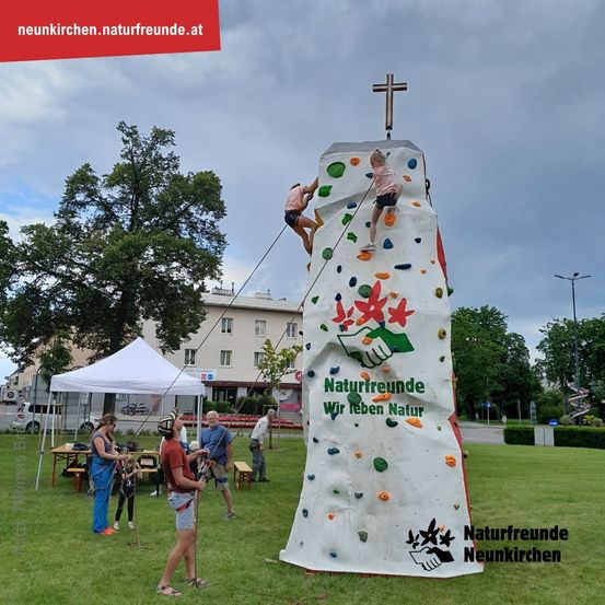Menschen klettern auf einer künstlichen Kletterwand, gesponsert von Naturfreunde, in einem Park. Zwei Kinder klettern, während andere zusehen.