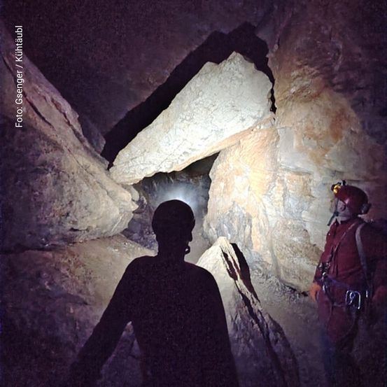 Zwei Personen erkunden eine Höhle mit Stirnlampen, eine steht neben einem großen Felsen.
