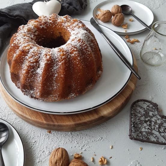 Ein Kuchen auf einem weißen Teller mit einem silbernen Messer, umgeben von Walnüssen, einer herzförmigen Dekoration und einem Glas.