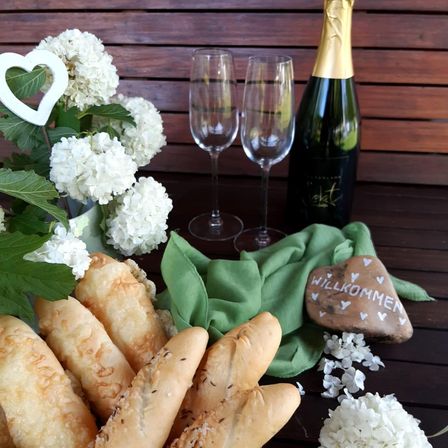 Ein Holztisch zeigt einen Strauß weißer Blumen, eine Flasche Wein, zwei Weingläser, einen herzförmigen Stein mit 'Willkommen' und eine Portion Brotstangen.