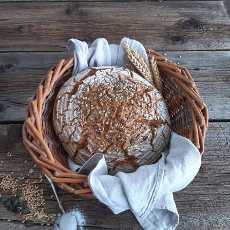Auf einem rustikalen Holztisch steht ein Korb mit einem Vollkornbrot, das mit Samen bedeckt ist, zusammen mit verstreuten Körnern und einem Tuch.