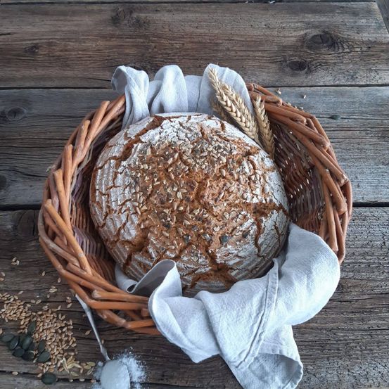 Auf einem rustikalen Holztisch steht ein Korb mit einem Vollkornbrot, das mit Samen bedeckt ist, zusammen mit verstreuten Körnern und einem Tuch.