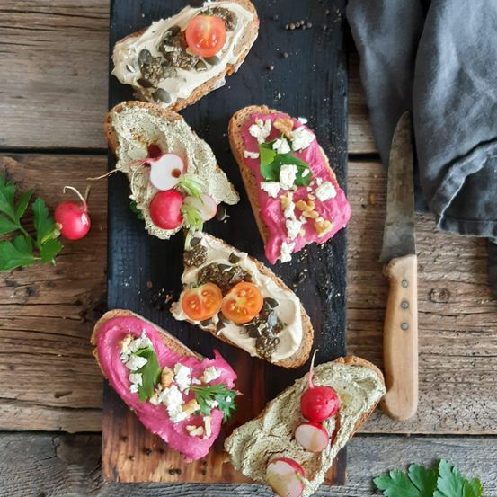 Ein Holzschneidebrett mit fünf Scheiben Brot, jede mit verschiedenen Belägen wie Radieschen, Tomaten und Kräutern. Ein Messer ist rechts.