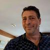 Ein lächelnder Mann macht ein Selfie und trägt ein blaues Hemd mit Gucci-Logo. Er steht in einem Raum mit weißen Wänden und einer hölzernen Decke.