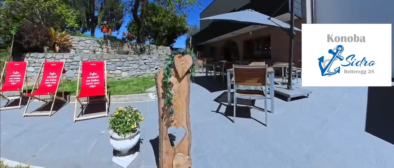 Eine Außensitzfläche eines Restaurants mit einem Baumstammpfosten, einem Topfpflanzen und einem Tisch mit Stühlen. Hinter dem Restaurant befindet sich eine Steinmauer und ein Baum.
