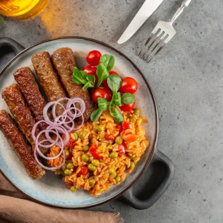Ein Teller mit Würstchen, Reis, Erbsen und Tomaten auf grauem Hintergrund mit Besteck.