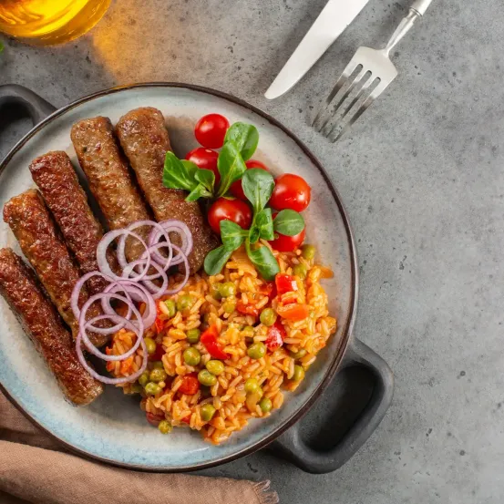 Ein Teller mit Würstchen, Reis, Erbsen und Tomaten auf grauem Hintergrund mit Besteck.