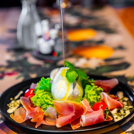 Ein Gericht mit einem halben Ei, Basilikum und Prosciutto auf einem Teller, garniert mit Kirschtomaten und grünem Gemüse. Im Hintergrund ein Tisch mit verschwommenen Gewürzen.