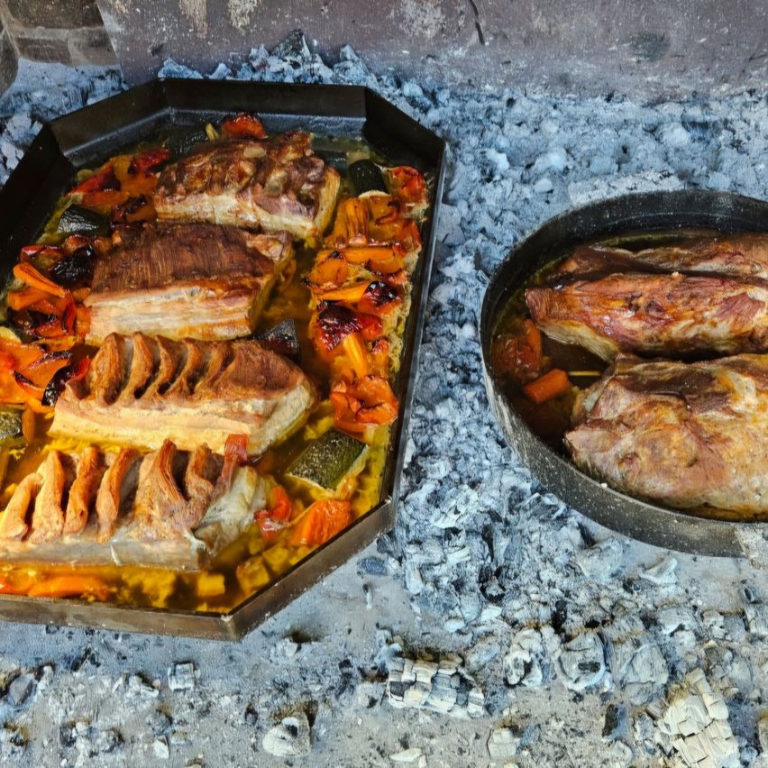 Two pans with cooked meats are placed on ashes. The meats are sliced and covered with a sauce. The ashes are scattered on the ground.
