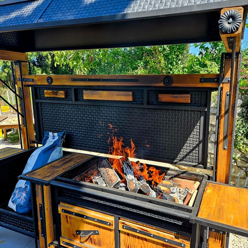 An outdoor grill setup with a wooden frame, metal roof, and a fire burning inside, surrounded by bags and trees.