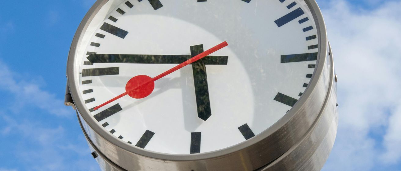 Eine große analoge Uhr mit einer roten Sekundenzeiger ist an einem Metallpfahl gegen einen blauen Himmel montiert.