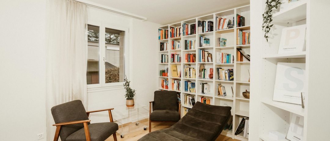 Eine gemütliche Leseecke mit einem Chaiselongue, zwei Stühlen und einem hohen Bücherregal voller Bücher. Ein Topf mit Pflanze steht auf dem Tisch zwischen den Stühlen. Der Raum hat Holzböden und weiße Wände.