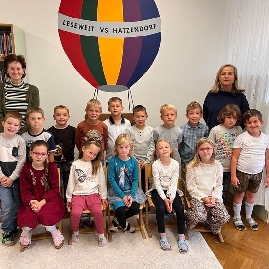 Eine Gruppe von Kindern und zwei Frauen posiert für ein Foto in einem Klassenzimmer mit einem farbenfrohen Heißluftballon im Hintergrund.