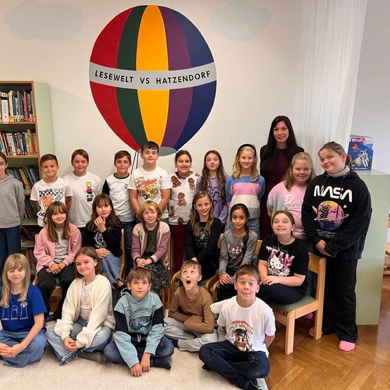 Eine Gruppe von Kindern und Erwachsenen in einem Klassenzimmer, posiert für ein Foto vor einer farbenfrohen Heißluftballon-Wandmalerei.