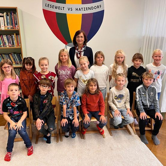Eine Gruppe von Kindern und einer erwachsenen Frau, die für ein Foto in einem Raum mit einem Heißluftballon-Logo an der Wand posiert.