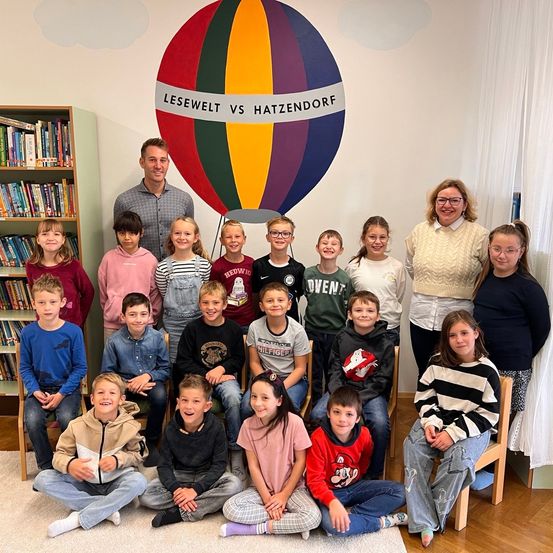 Eine Gruppe von Kindern und Erwachsenen in einem Raum mit einem farbenfrohen Heißluftballon-Wandbild. Einige sitzen auf Stühlen, andere stehen dahinter und lächeln zur Kamera.