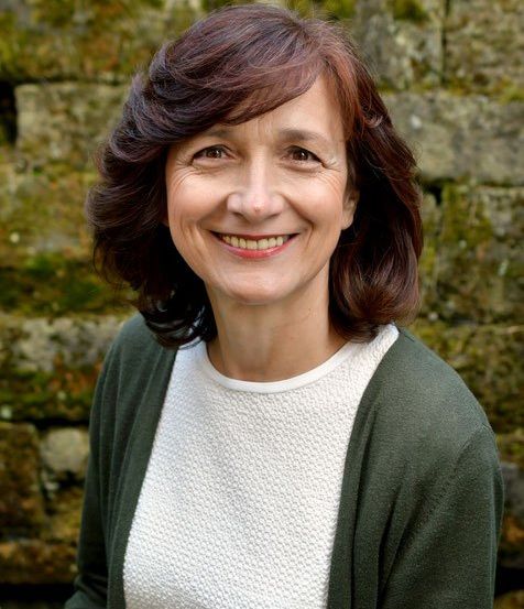 Eine Frau mit kurzen braunen Haaren und einem weißen Top lächelt vor einer moosbewachsenen Steinmauer.