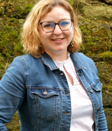 Eine Frau mit blonden Haaren, die eine Brille und eine Jeansjacke trägt, lächelt vor einer moosbewachsenen Felswand.