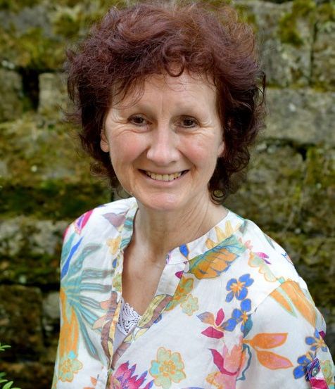Eine Frau mit kurzen, lockigen, braunen Haaren und einem Blumenhemd lächelt vor einer mit Moos bedeckten Steinmauer.