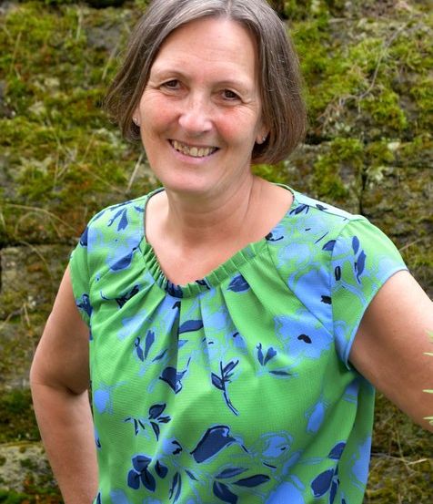 Eine Frau steht vor einer mit Moos bedeckten Steinmauer. Sie lächelt und trägt ein grünes und blaues Blumenhemd.