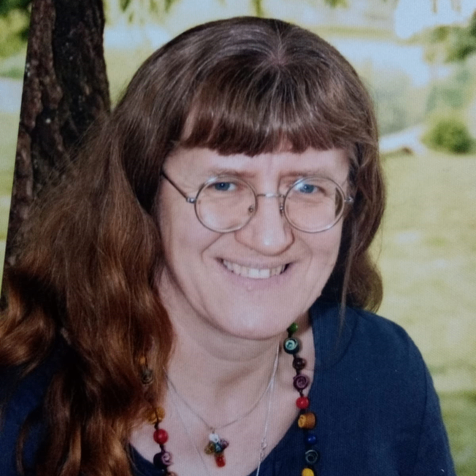 Eine Frau mit braunem Haar und Brille lächelt im Freien. Sie trägt ein blaues Shirt und eine bunte Halskette. Hinter ihr ein Baumstamm und ein verschwommener Hintergrund.
