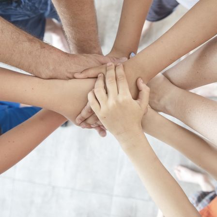 Mehrere Menschen unterschiedlichen Alters halten in einem Kreis die Hände, möglicherweise als Symbol für Einheit oder Unterstützung.