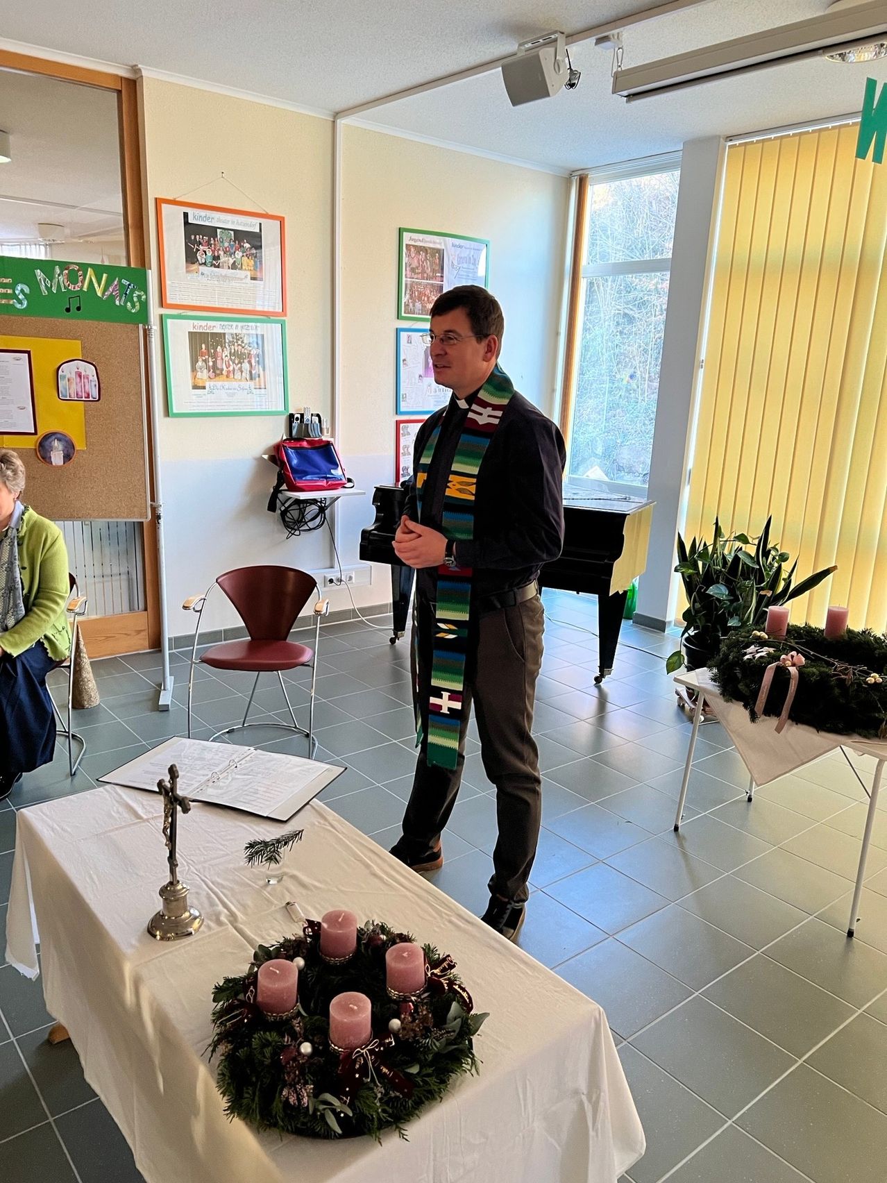 Ein Mann steht neben einem Tisch mit einem Kranz aus Kerzen in einem Raum mit einem Klavier, Stühlen und einer Anschlagtafel.