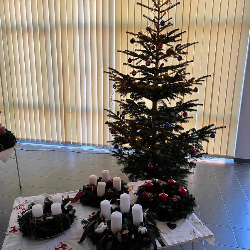 Ein geschmückter Weihnachtsbaum mit bunten Kugeln und Lichtern steht. Zwei Kränze mit Kerzen sind auf einem Tisch davor. Der Raum hat geflieste Böden und vertikale Jalousien.