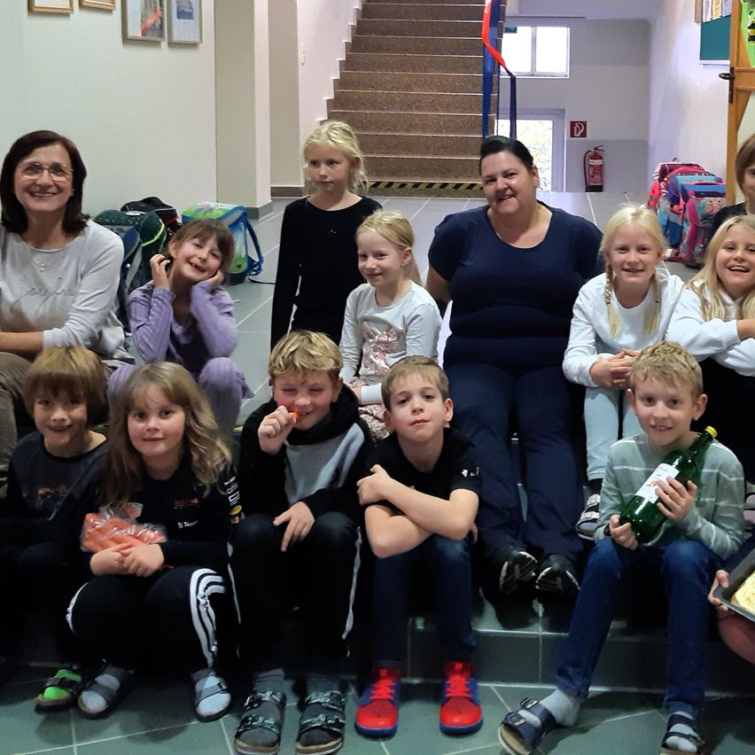 Eine Gruppe von Kindern und Erwachsenen posiert für ein Foto in einem Raum, einige halten Snacks und Getränke, mit einer Treppe und gerahmten Bildern im Hintergrund.