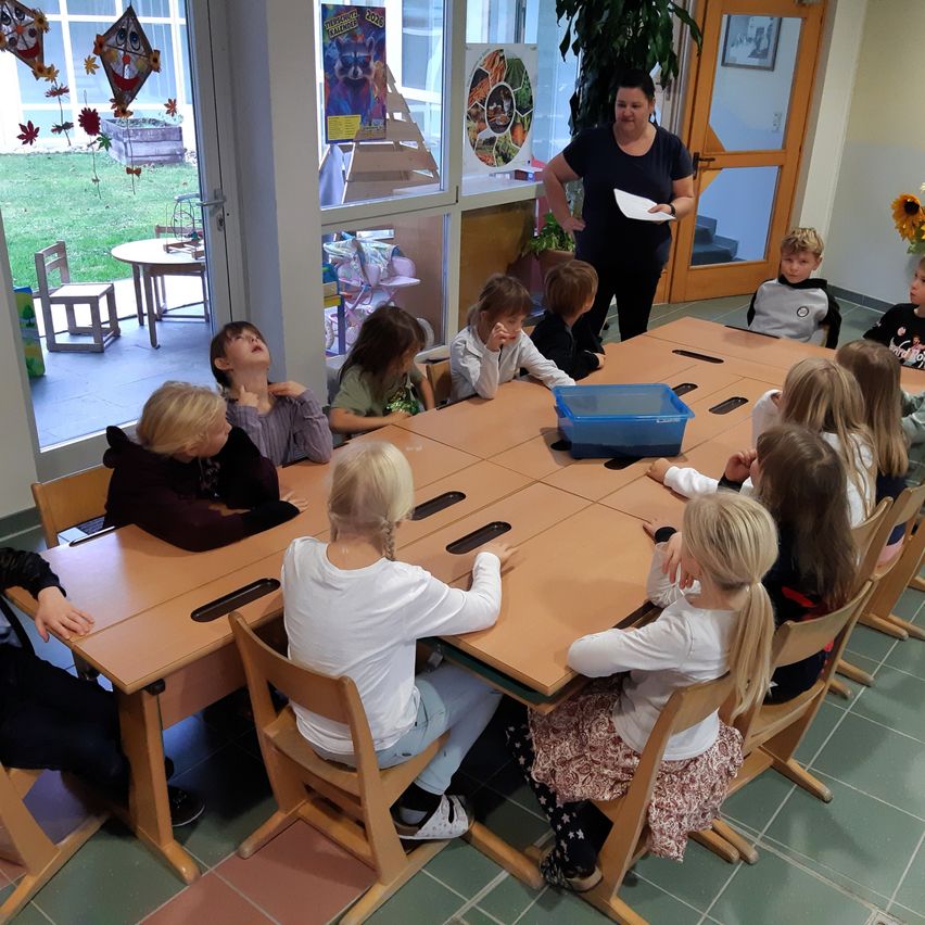 Eine Gruppe von Kindern sitzt an einem Tisch in einem Klassenzimmer. Eine Frau steht vorne und hält ein Papier. Das Klassenzimmer hat Glastüren, Stühle und einen Tisch mit einer blauen Aufbewahrungsbox. Draußen gibt es einen Rasenbereich und eine Holztür.