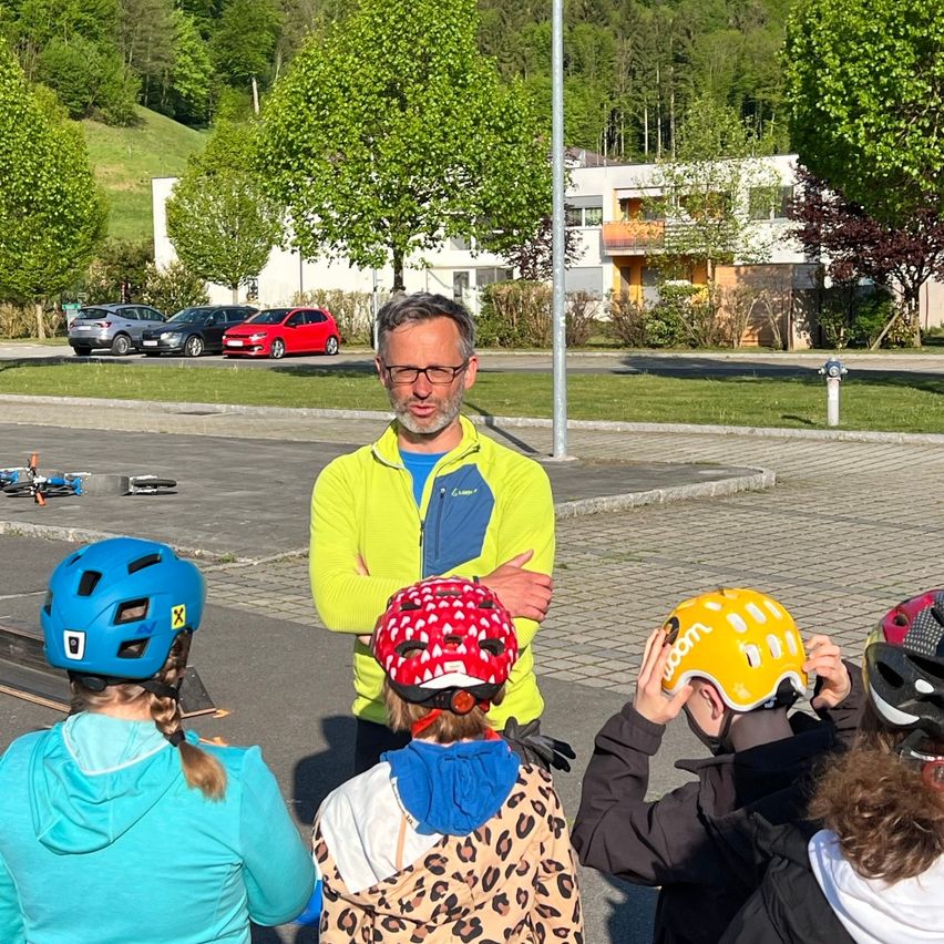 Ein Mann mit Brille steht mit verschränkten Armen neben Kindern, die Helme tragen. Sie sind draußen in der Nähe eines Gebäudes und einiger geparkter Autos.