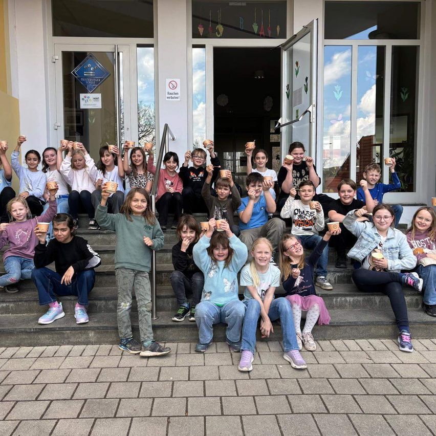 Eine Gruppe von Kindern, die Becher mit Essen halten, stehen auf den Stufen eines Gebäudes mit Glastüren und Fenstern. Einige halten ihre Hände in der Luft.