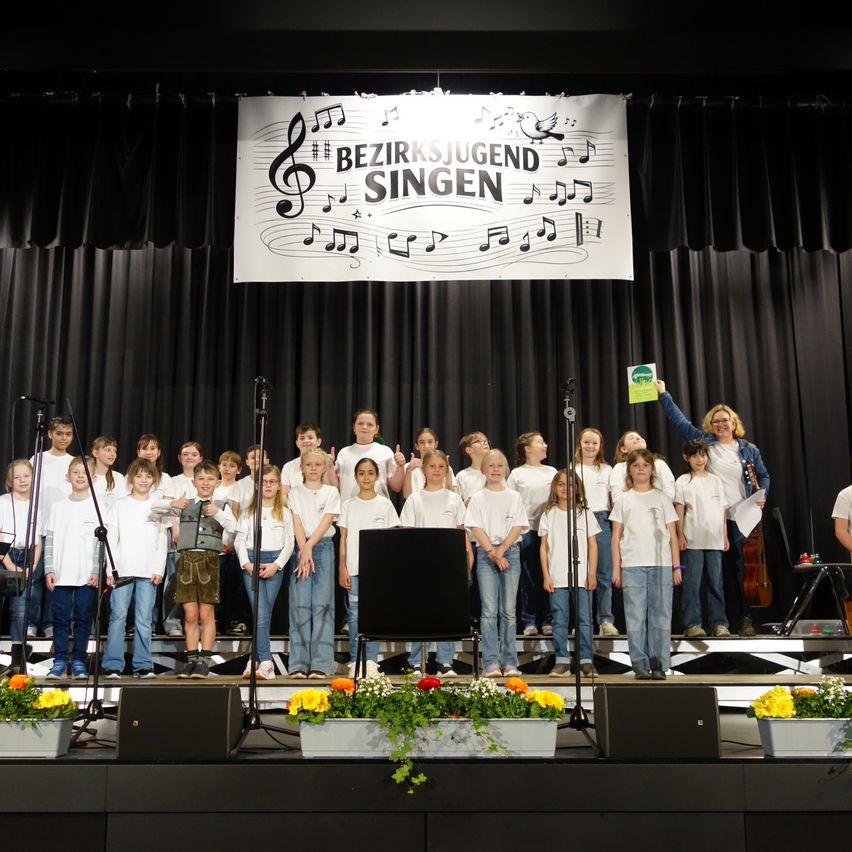 Eine Gruppe von Kindern in weißen T-Shirts steht mit Mikrofonen auf der Bühne und singt, während eine Frau ein Buch hält. Dahinter befindet sich eine Bühnenkulisse mit Noten und einem Banner.
