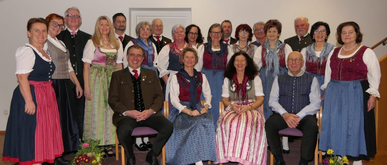 Eine Gruppe von Menschen in traditioneller Kleidung posiert für ein Foto. Einige sitzen, andere stehen hinter ihnen. Sie tragen alle festliche Kleidung mit farbenfrohen Details. Ein Blumenarrangement steht auf dem Boden davor.