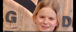 Eine Frau mit blondem Haar steht vor einem hölzernen Hintergrund mit dem Text 'Wir dürfen euch allen heute unseren Lehrling Anja Lang vorstellen.' Sie trägt ein schwarzes Hemd.