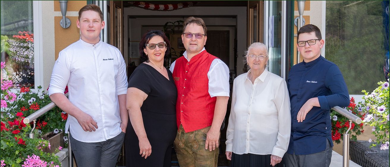 Vier Personen stehen vor einer Tür. Ein Mann und eine Frau tragen traditionelle Kleidung. Die Frau auf der linken Seite trägt ein schwarzes Kleid. Die andere Frau trägt ein weißes Hemd.