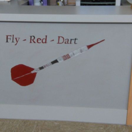 Eine weiße Wand mit rotem Text 'Fly - Red - Dart' und einem roten Pfeil mit roter Spitze und silbernem Schaft, der daran befestigt ist.