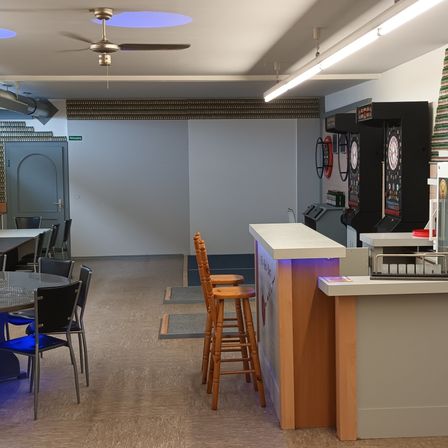 Ein Raum mit Deckenventilator, Beleuchtung und Dartscheibe. Es gibt Stühle und Tische sowie eine Theke mit Registrierkasse. Der Raum hat einen Holzboden.