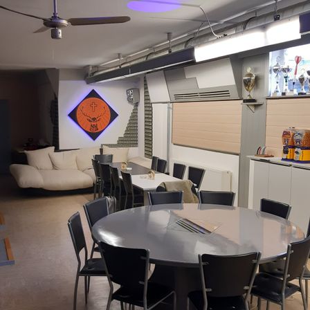 Ein Indoor-Essbereich mit einem großen runden Tisch, umgeben von Stühlen, einer Couch und einer Wand mit Hirsch- und Kreuzdesign. Oben befindet sich ein Deckenventilator, und Trophäen stehen auf einem Regal.
