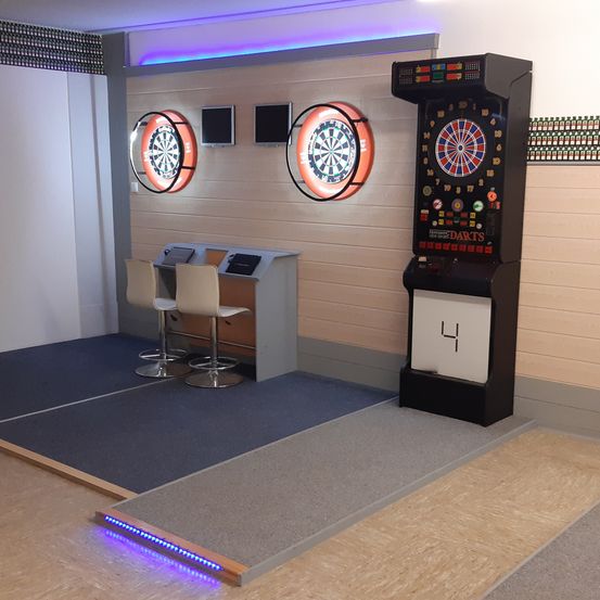 Ein Dartraum mit zwei Dartscheiben an der Wand, einem schwarzen Dartautomaten, zwei weißen Stühlen und blauer Beleuchtung auf dem Boden.