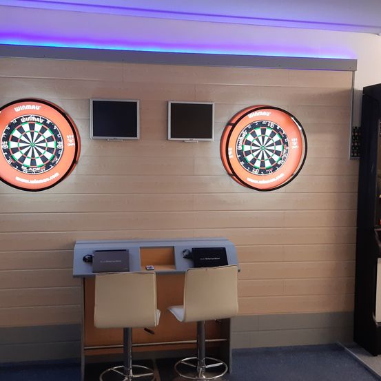 Zwei Dartscheiben hängen an einer Holzwand mit zwei Monitoren darüber. Ein Dartboard ist auch auf jeder Seite des Raumes ausgestellt. Zwei Hocker sitzen vor einer Theke.