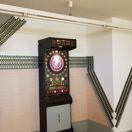 Ein Dartautomat ist in einer Ecke eines Raumes mit weißen Wänden installiert. Die Wände haben Dartscheiben. Eine weiße Röhre ist an der Decke befestigt.