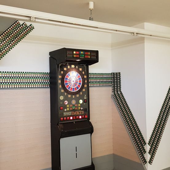 Ein Dartautomat ist in einer Ecke eines Raumes mit weißen Wänden installiert. Die Wände haben Dartscheiben. Eine weiße Röhre ist an der Decke befestigt.